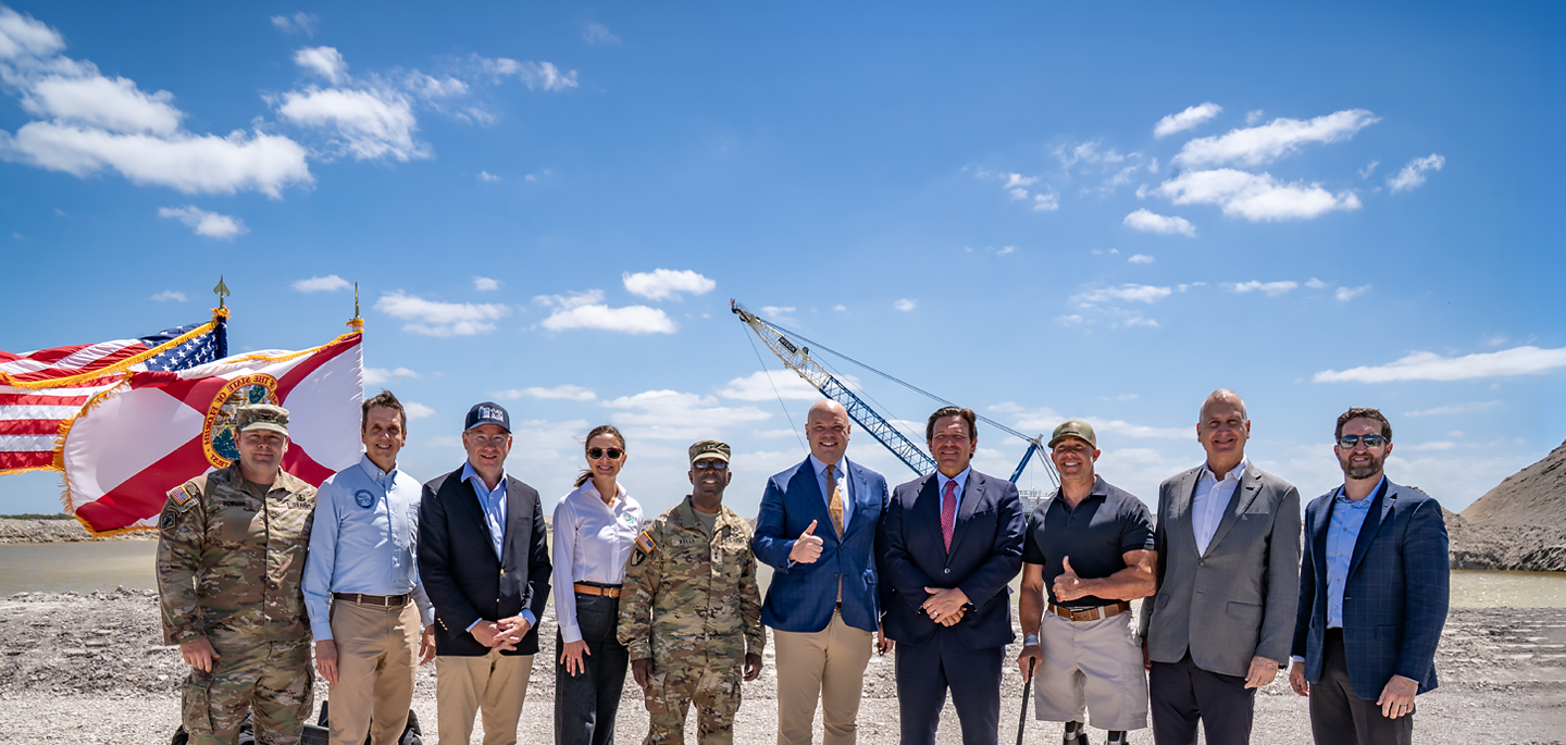 State and Federal Officials stands together in front of flags and a large crane at the EAA Reservoir